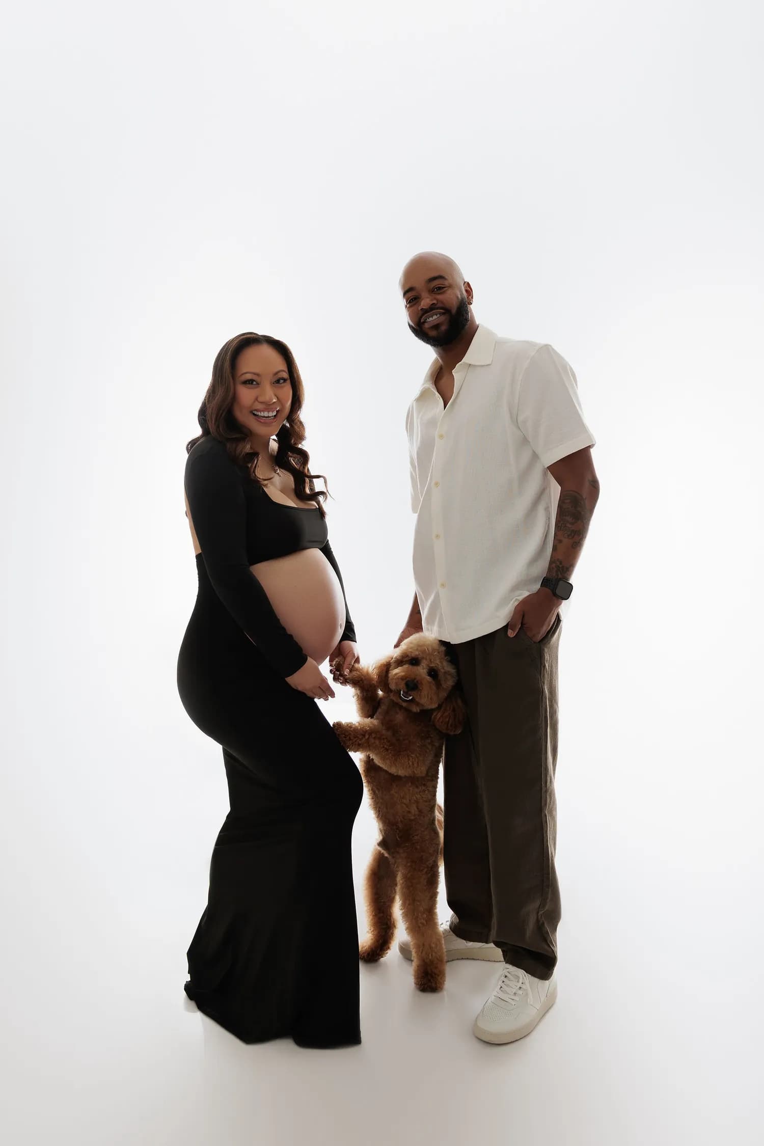 An expecting couple on a white background with a curly-haired dog standing up against her