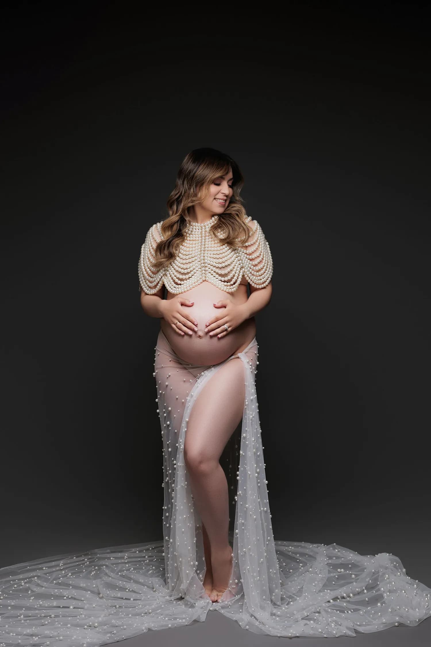 Pregnant woman in a pearl cape and sheer pearl skirt against a dark backdrop