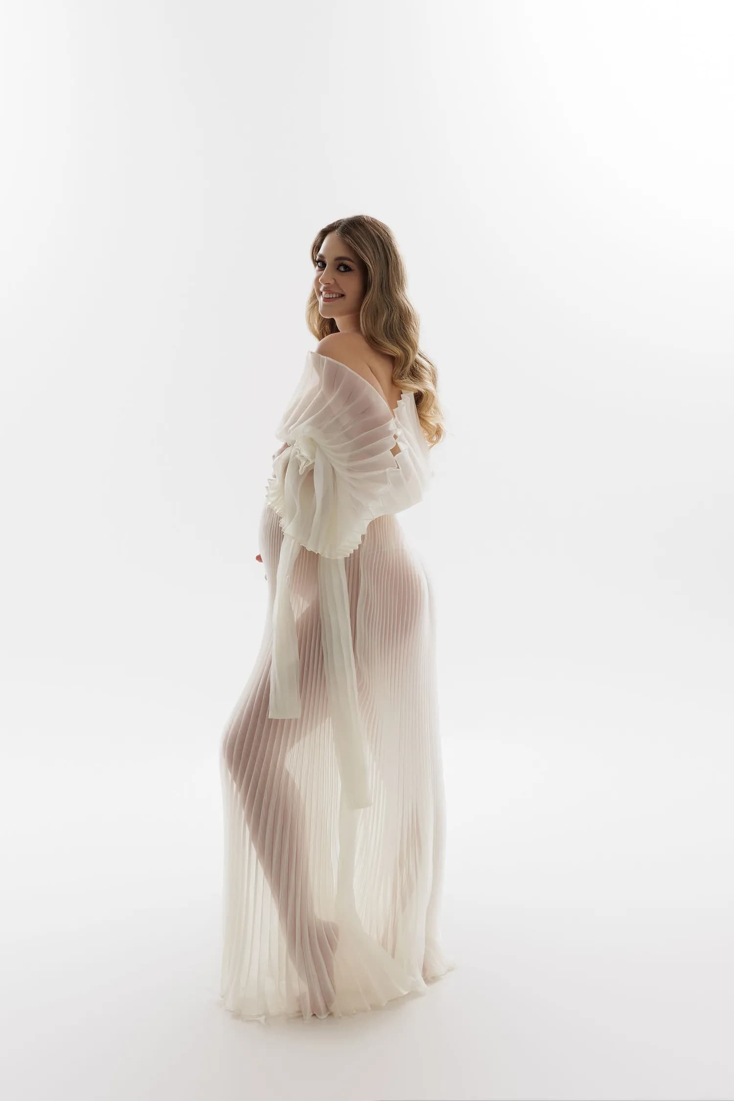 Pregnant woman in a sheer ivory pleated gown, smiling over her shoulder