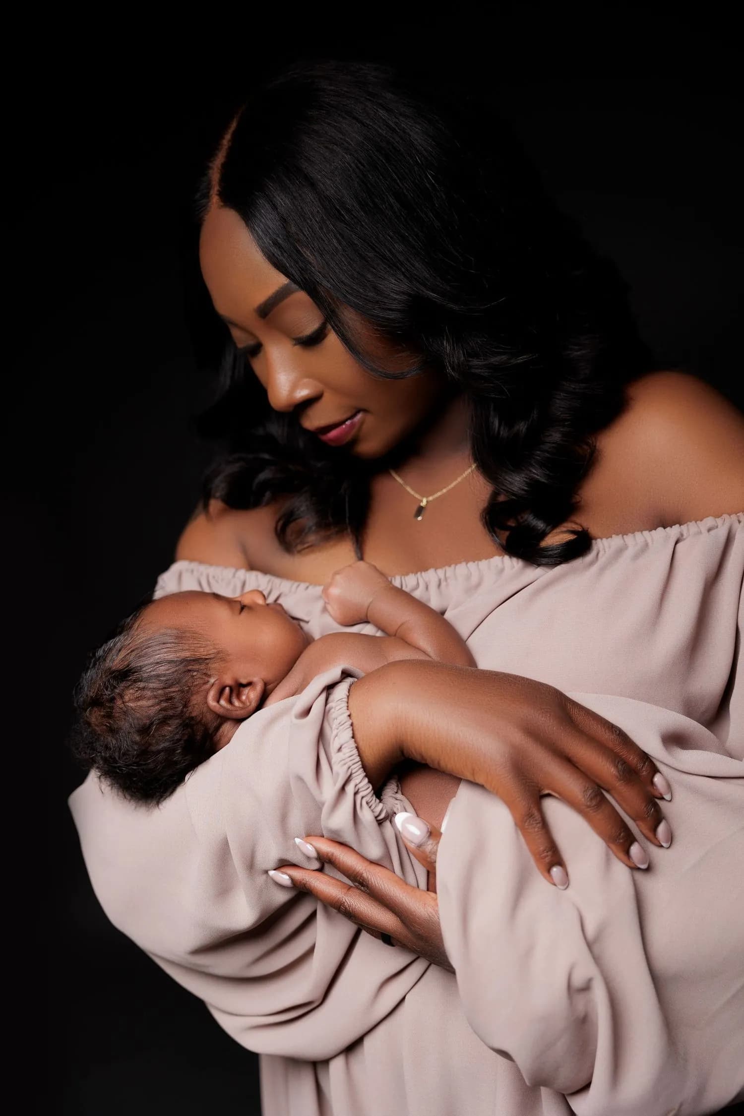 A mother in an off-the-shoulder taupe dress looks down at the sleeping newborn she cradles against her chest