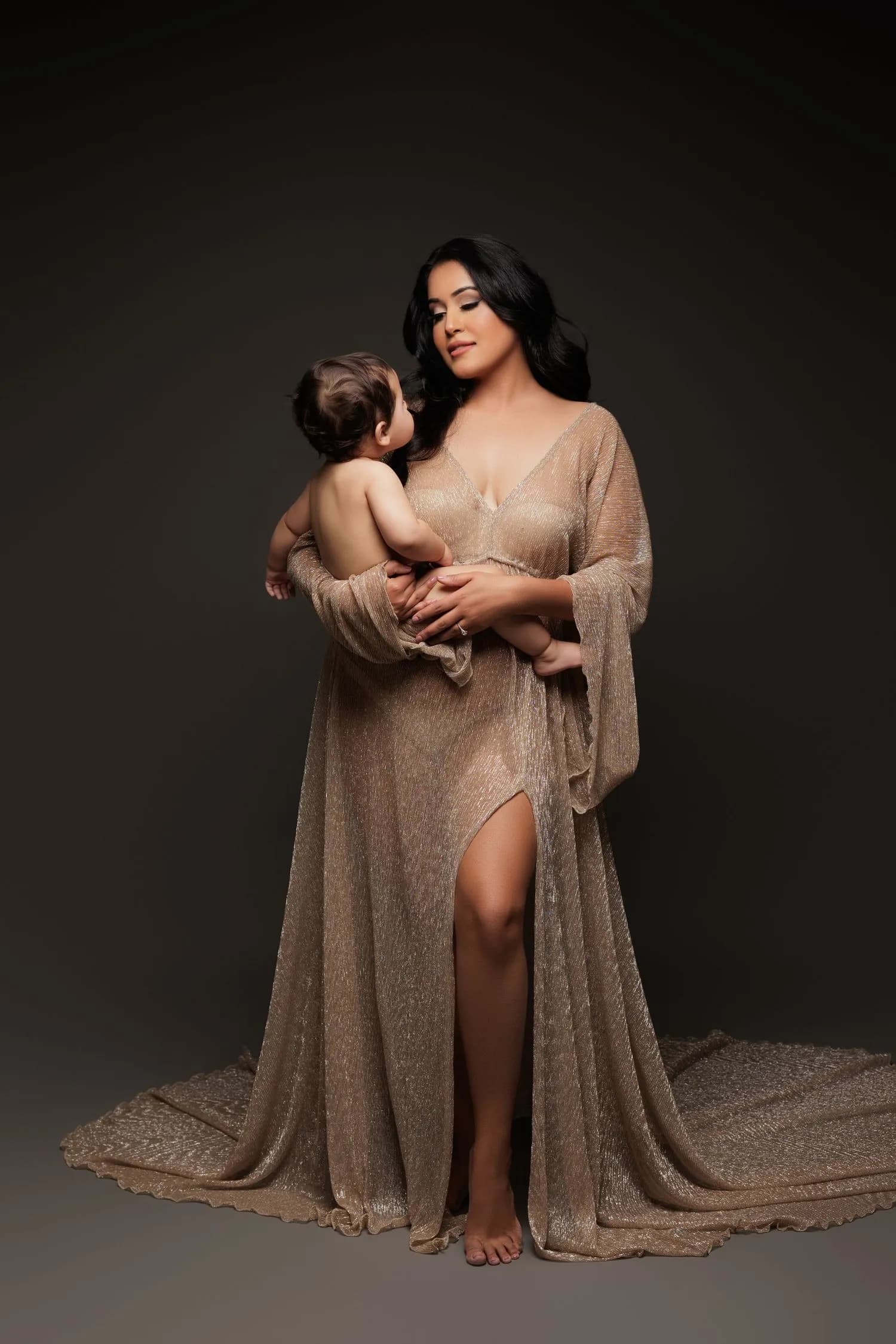 A mother in a long shimmering gold gown with a flowing train stands barefoot against a dark backdrop, holding her baby, who looks up at her